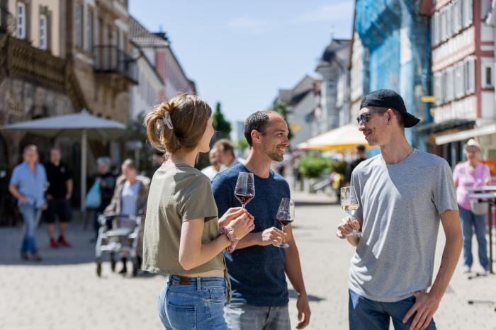 Drei junge Leute stehen mit einem Weinglas in der Hand in der Fußgängerzone. 