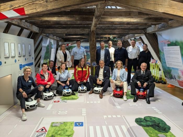 Die Teilnehmer der Ausstellungseröffnung sitzen auf Bobby Cars. 