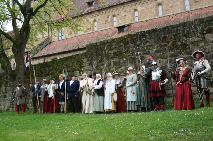 Darsteller in historischen Kostümen stehen vor einer Stadtmauer. 