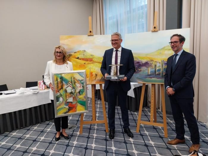 Drei Personen stehen mit Geschenken nebeneinander. Die Geschenke sind drei Gemälde und eine Skulptur.