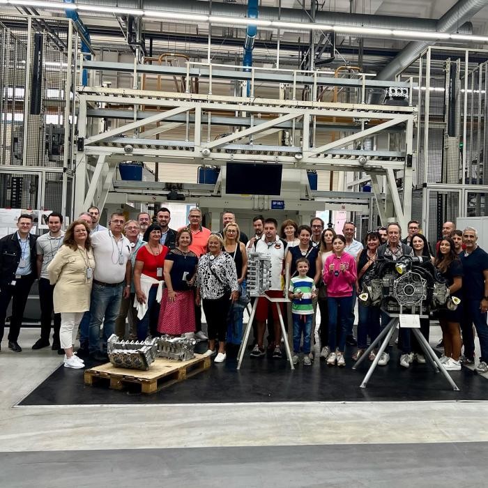 Gruppenfoto bei der Führung durch die Gießerei der KS HUAYU AluTech GmbH Eine Gruppe von Menschen steht hinter einem Automobilmotor.