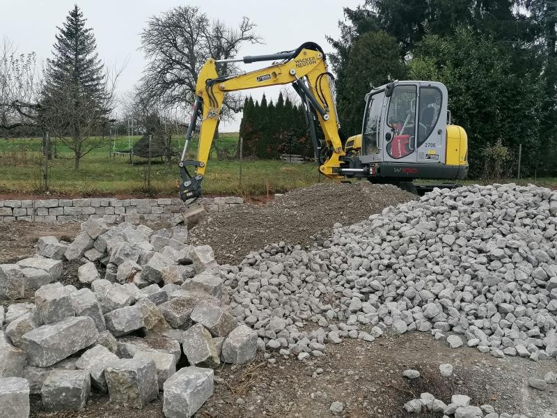 Auf dem Schotterparkplatz arbeitet ein Bagger am Bau der Trockenmauer.