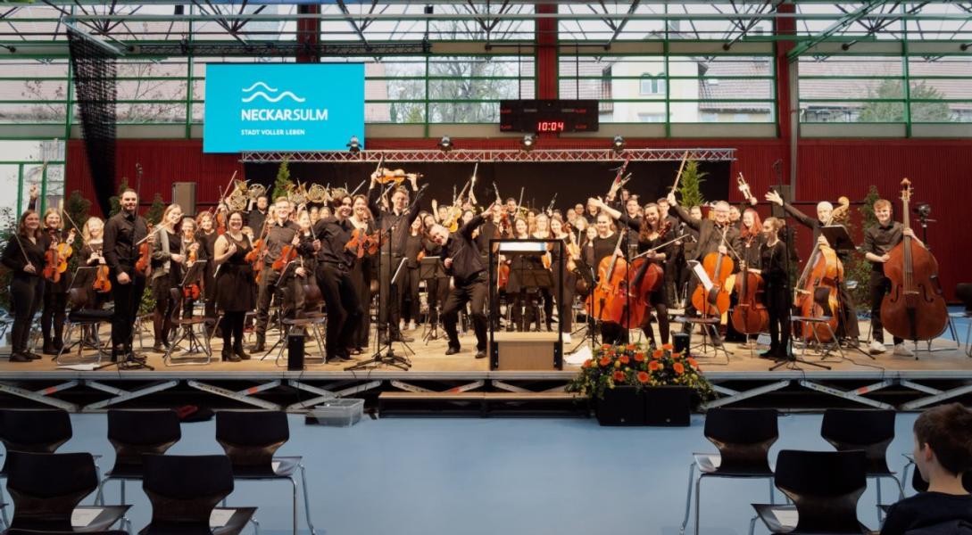 Das Jugend-Sinfonieorchester steht auf der Bühne in der Ballei-Sporthalle und winkt in die Kamera.