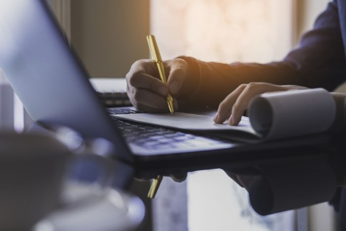 Foto: Adobe Stock Eine Person hält einen Stift und macht sich Notizen auf einem Papier. Davor steht ein Laptop.