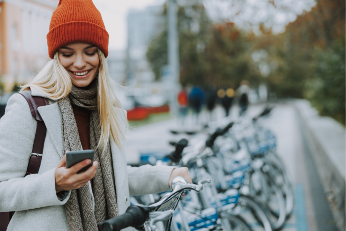 Eine Frau hält ein Smartphone in der Hand und schaut auf das Display. Im Hinterrund sind Fahrräder zu sehen.