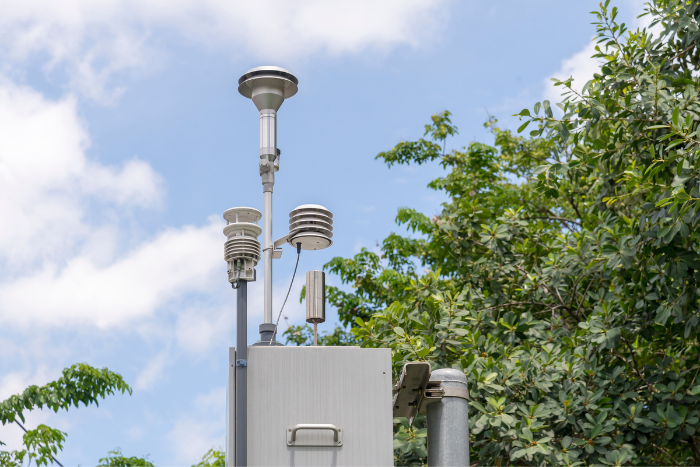 Eine Luftqualität-Messstation steht vor grünen Bäumen.